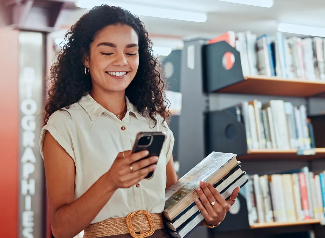 A smiling woman with curly hair holds a stack of books and a smartphone in a library aisle.