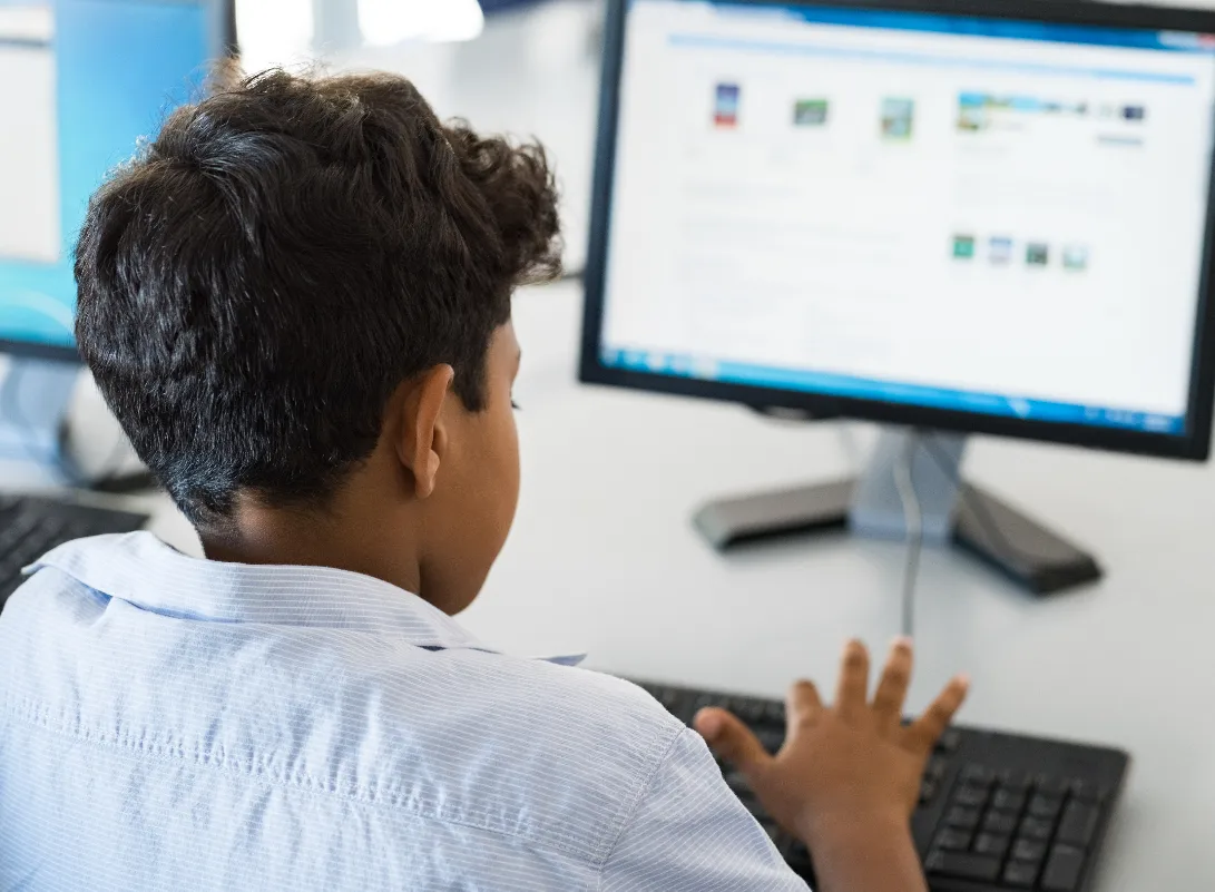A young boy in a light blue collared shirt uses a computer, his hands on the keyboard and eyes focused on the screen.