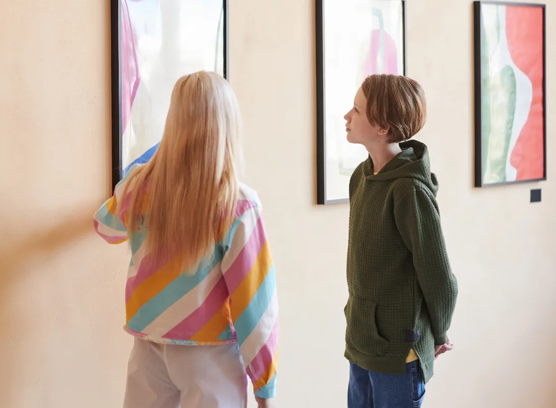 Two children, a girl with blonde hair and a boy, observe abstract art in a gallery.