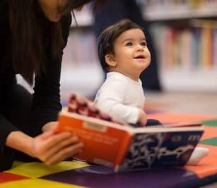 A smiling baby looks up while an adult reads a book to them, surrounded by colorful mats and bookshelves.
