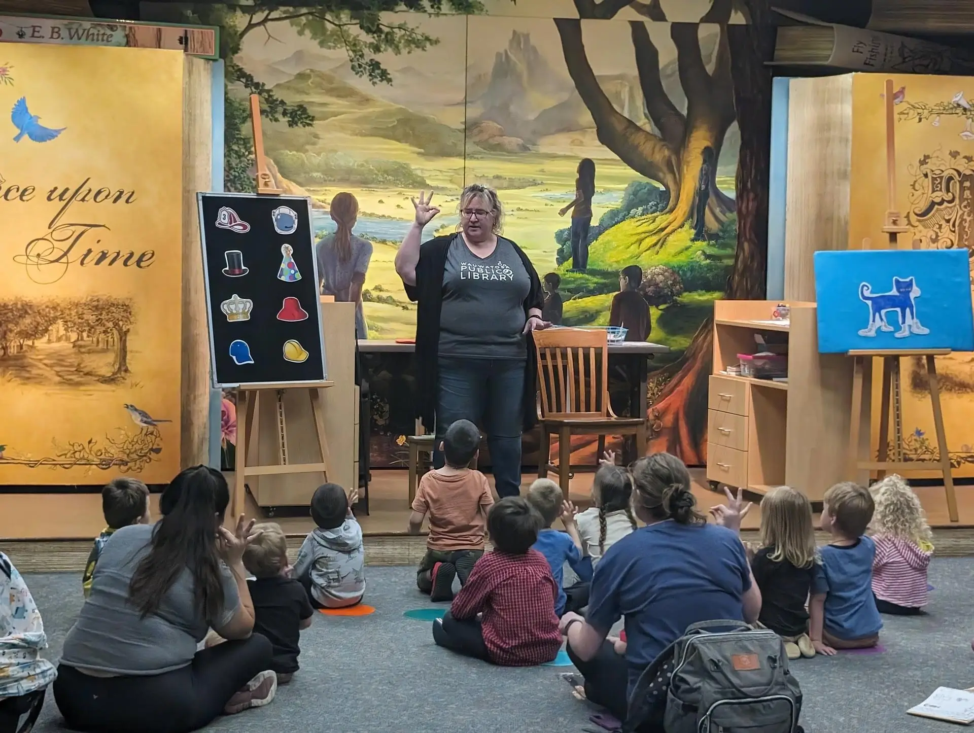 A librarian leads a Rhyme Time program for young children, using props like hats and a blue cat cutout.