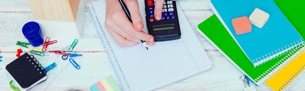 A person's hands using a calculator and pencil on a grid notebook, surrounded by school supplies like notebooks, erasers,