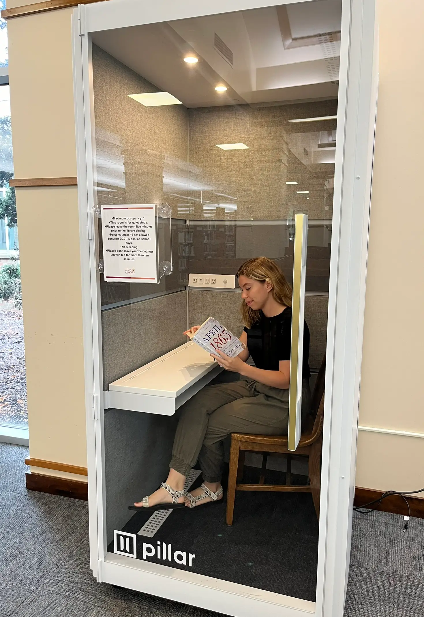 A young woman sits in a Pillar quiet study booth, reading a book titled "April 1865".