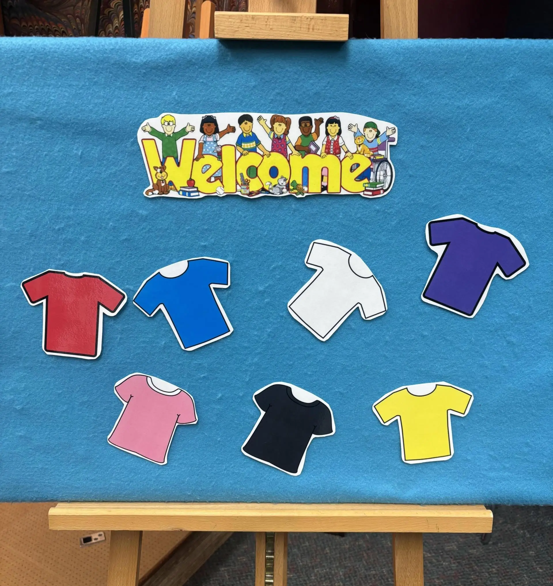 A "Welcome" sign with diverse cartoon children and a dog, above seven colorful t-shirt cutouts on a blue felt board.