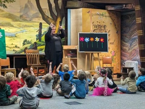 A librarian leads a story time session for children, using hand gestures and a colorful flower display.